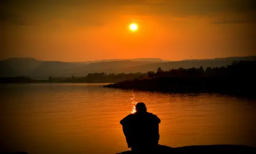 An underwhelmed man sitting by the river and watching the sunset